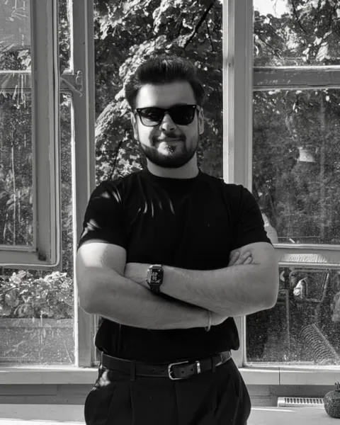 Osman Yağmur, siyah tişört ve güneş gözlüğüyle, kolları kavuşturulmuş halde açık pencerelerin önünde duruyor. Siyah beyaz fotoğraf.