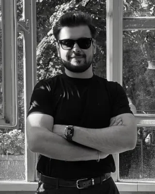 Osman Yağmur, siyah tişörtle, kolları kavuşturulmuş halde açık pencerelerin önünde duruyor. Siyah beyaz fotoğraf.