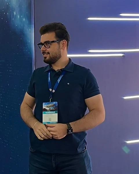 Osman Yağmur, gözlüklü ve lacivert polo tişörtlü, yarışmacı kartıyla Teknofest Finansal Teknolojiler Yarışması alanında duruyor.